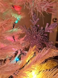 close up of a snowflake shaped oranment on a white christmas tree with colored lights around 