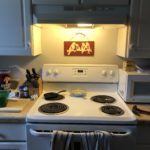 Electric Kitchen stove with four burners. Light above the stove is on there is a red wooden plaque with the silhouette of four birds and the word love