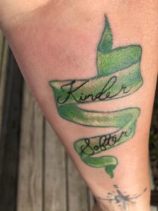 Tattoo of green ribbon with two words Kinder and Softer written on it