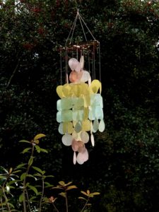 pastel colored pink red, yellow, blue glass wind chimes haning in front of tall green bushes and berries.
