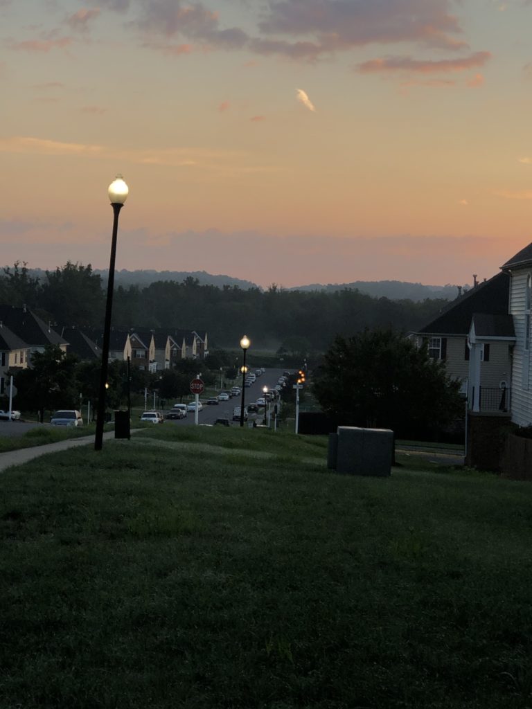 a dark street, looking across a neighborhood to hills with a sky lightening from purple to orange as the sun comes up