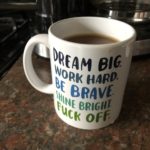 A regular sized white coffee mug, with series of motivational phrases written in blue and green text from the top to the bottom. Dream Big, Work Hard, Be Brave, Shine Bright, Fuck Off