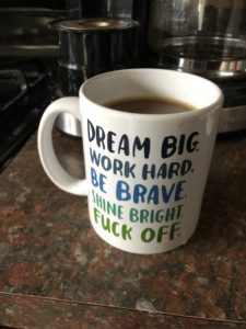A regular sized white coffee mug, with series of motivational phrases written in blue and green text from the top to the bottom. Dream Big, Work Hard, Be Brave, Shine Bright, Fuck Off