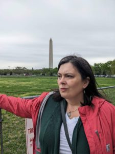 A woman in a pink coat stands with her hand on a fence looking into the distance with a small scowl and furrowed brow. The Washington Monument is in the background.