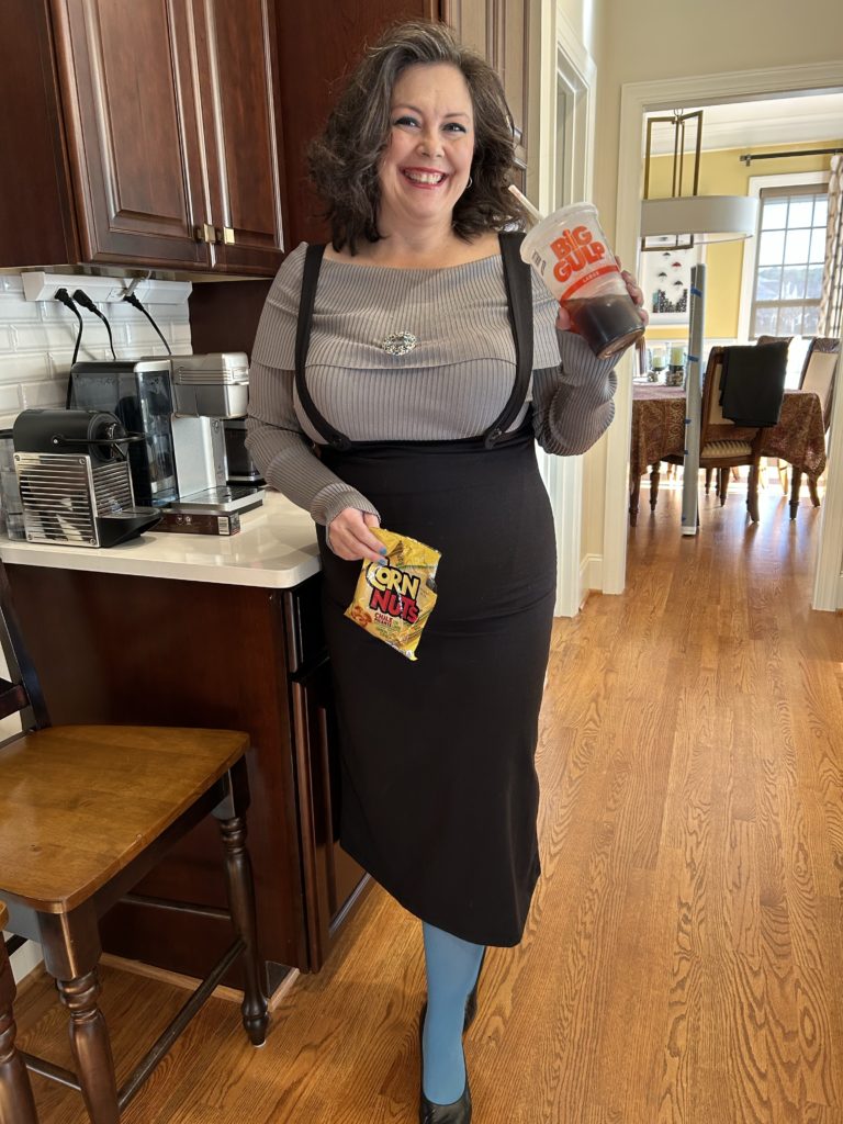 A woman stands in a grey off the shoulder top with a black bib jumper with a mid calf lenght skirt. She has on blue tights, and is holding a large gas station soda and a bag of Corn Nuts. 