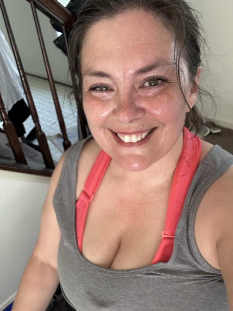 A visibly sweaty, smiling woman with hair pulled back in a pony tail wears a scoop neck grey tank top and an orange sports bra.