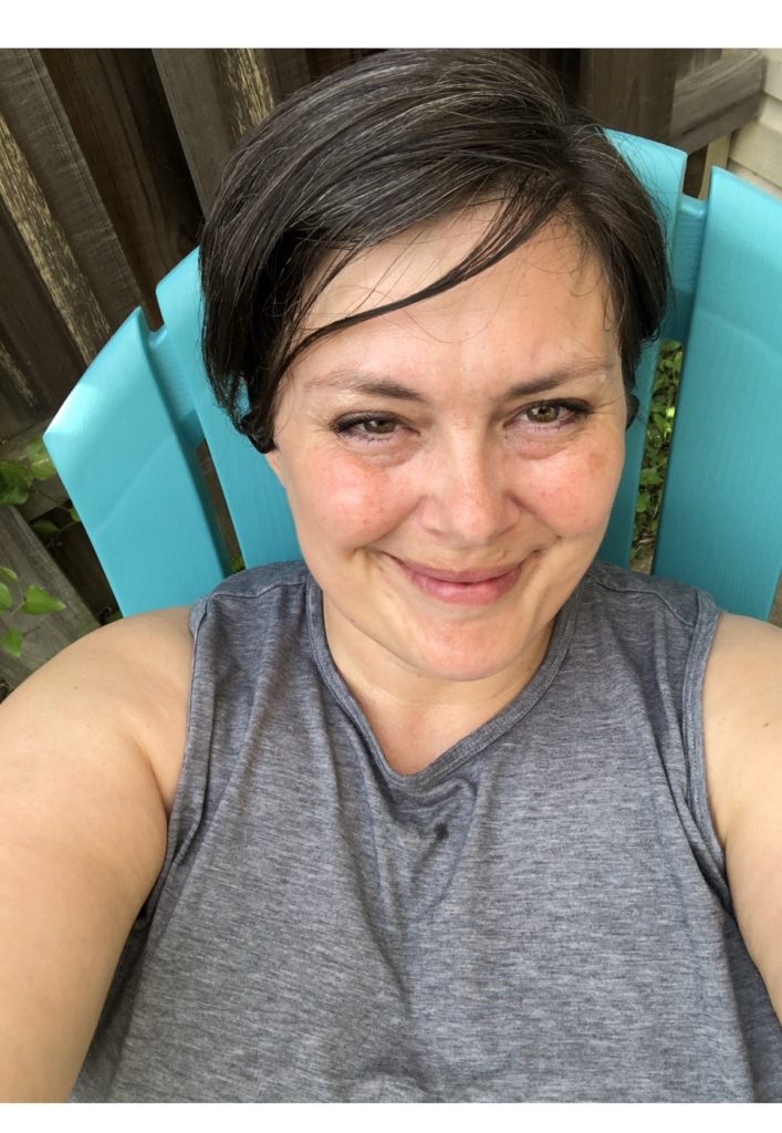 April 2021. A smiling woman with short hair, bangs sweeping across her forehead, wears a high neck grey tank top and sits in a teal Adirondack style lawn chair. 