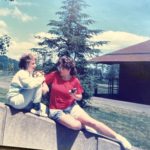 On a sunny day, two young girls sit on a sloped retaining wall. One has her knees pulled up and is smiling and talking. The other is turned, her full body to the camera, leaning on her right hip, her left leg outstretched, face turned to her companion. She is holding a small 35mm camera in her hand.