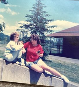 On a sunny day, two young girls sit on a sloped retaining wall. One has her knees pulled up and is smiling and talking. The other is turned, her full body to the camera, leaning on her right hip, her left leg outstretched, face turned to her companion. She is holding a small 35mm camera in her hand.