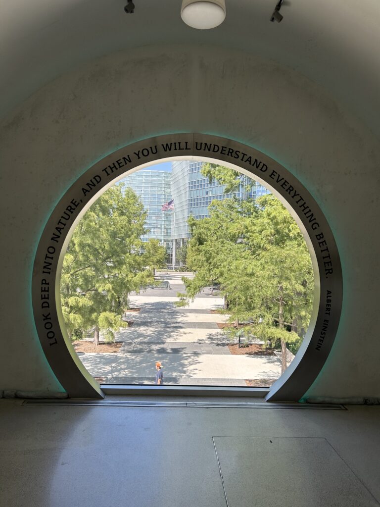 Circular window out onto a tree lined brick and concrete path.  A quotation carved in to the frame says, “Look deep into nature, and then you will understand everything better.” 