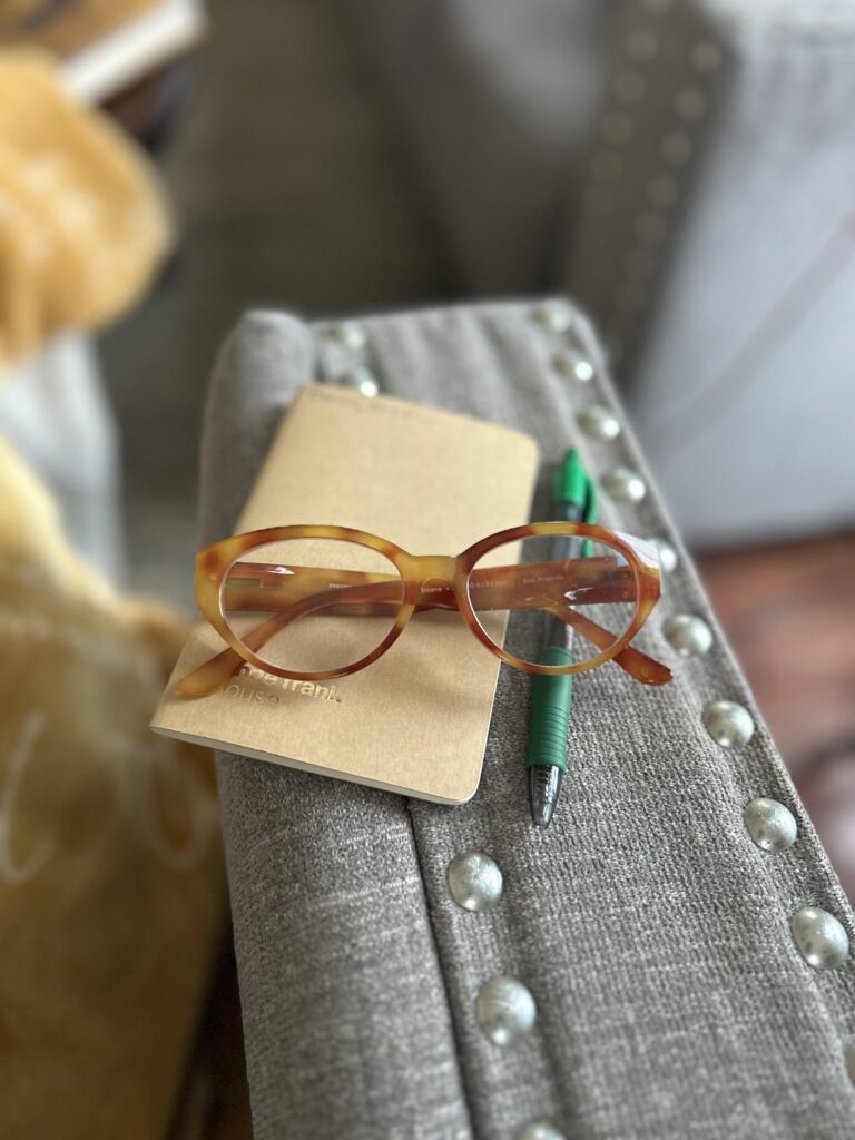 The flat rectangle of a grey couch arm. A small tan notebook from the Anne Frank House sits next to a green pen. Books 2025- is written in green ink at the top of the notebook cover. A pair of reading glasses with tan tortoise shell frames sits on top of the notebook and pen.